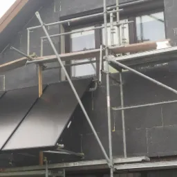 Dom w trakcie ocieplania: szara styropianowa elewacja, rusztowanie, okna z brązowymi ramami i panele słoneczne w dolnej części ściany.