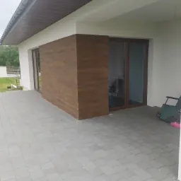 Wykończona elewacja domu z brązową okładziną imitującą drewno, widoczne duże przesuwne okna i szara kostka brukowa na tarasie
