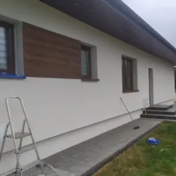 Świeżo pomalowana, biała elewacja domu jednorodzinnego z brązowymi oknami i panelem imitującym drewno, widoczna drabina malarska i resztki folii ochronnej na kostce brukowej.