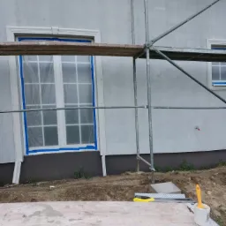 Świeżo otynkowana szara elewacja domu w trakcie prac wykończeniowych; okna zabezpieczone folią i taśmą, rusztowanie, narzędzia na podeście, widoczny fragment elewacji z ozdobnymi elementami.