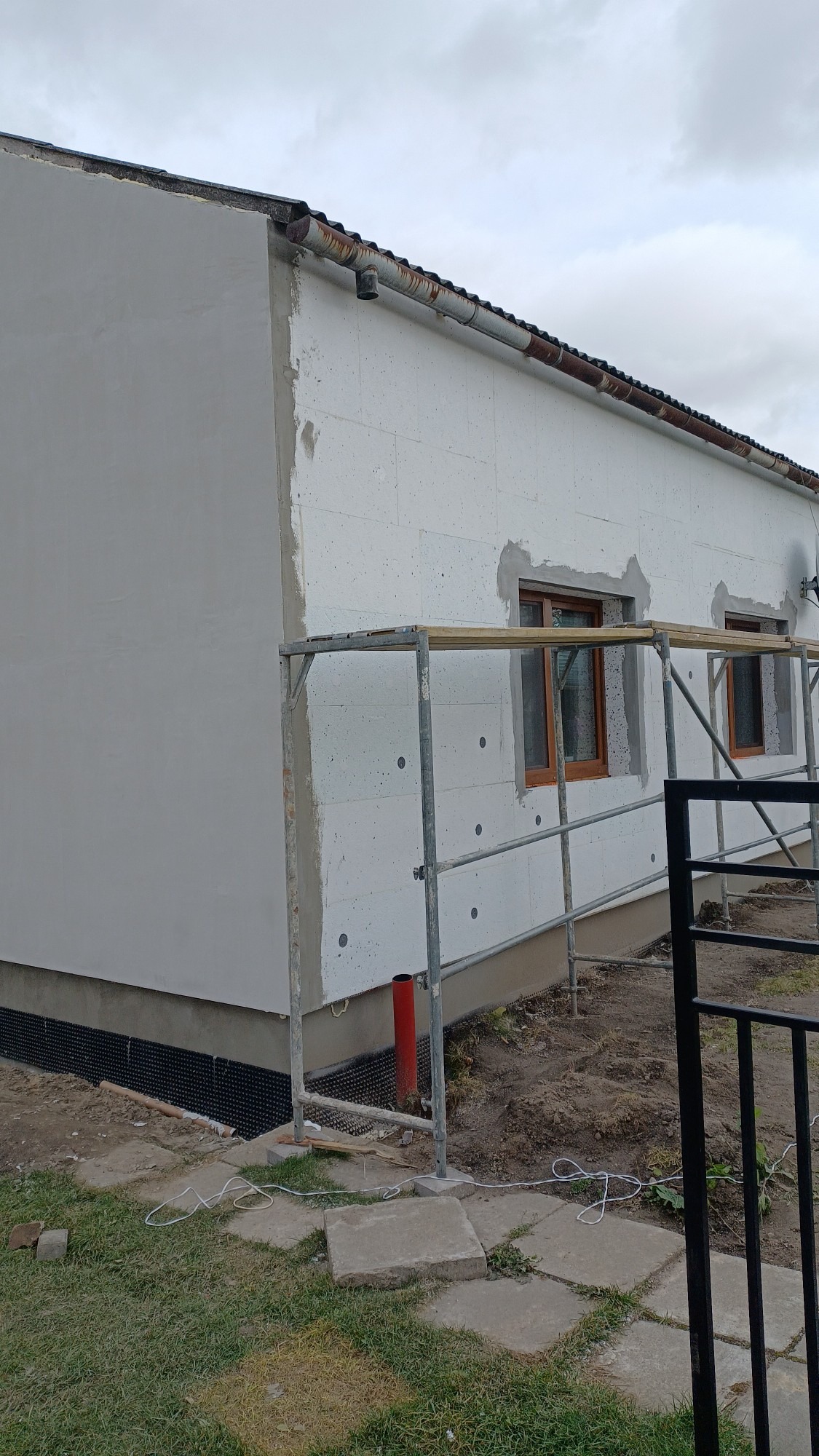 Dom jednorodzinny w trakcie ocieplania, widoczne fragmenty elewacji otynkowane i pokryte styropianem, rusztowanie, rynna z widoczną korozją, teren wokół domu częściowo zagospodarowany.