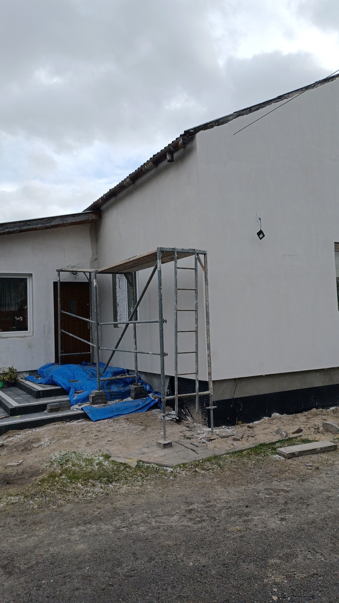 Dom jednorodzinny w trakcie prac elewacyjnych, widoczne rusztowanie przy wejściu, zabezpieczone folią schody i świeżo otynkowana, pomalowana na biało ściana budynku.