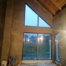 Wnętrze domu w trakcie prac wykończeniowych. Ściana szczytowa z dużym trójkątnym oknem i oknem przesuwnym poniżej, pokryta świeżym, surowym tynkiem. Widoczne drewniane elementy konstrukcji dachu.