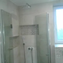 Nowoczesna kabina prysznicowa z deszczownicą i baterią termostatyczną, wykończona jasnymi płytkami z dekoracyjnymi wstawkami, oświetlona punktowo.