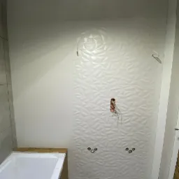 Łazienka w trakcie remontu: biała wanna z drewnianą obudową, ściana z trójwymiarowych białych płytek, widoczne instalacje hydrauliczne i elektryczne, fragment podłogi zabezpieczony folią.