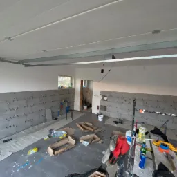 Wnętrze w trakcie układania szarych płytek na ścianie, widoczne krzyżyki dystansowe, narzędzia i materiały budowlane rozsypane na podłodze, przygotowane do fugowania.