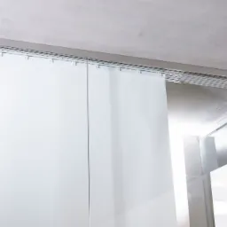 Minimalistyczne, białe rolety rzymskie zamontowane pod sufitem, widoczny mechanizm sterujący Somfy, jasne wnętrze.