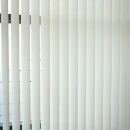 Zamknięte, kremowe żaluzje pionowe z widocznym fragmentem okna w tle.