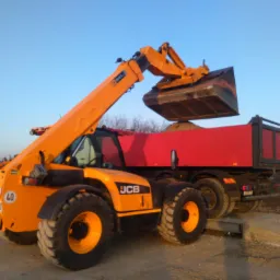 Pomarańczowa ładowarka teleskopowa JCB z pełną łyżką nad czerwoną wywrotką, przygotowująca się do załadunku ziemi lub gruzu na placu budowy.