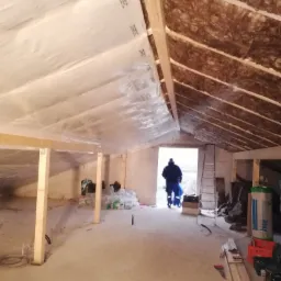 Poddasze w trakcie prac: izolacja wełną mineralną pod skosami dachu, folia paroizolacyjna na suficie, widoczny pracownik w oddali, drabina, materiały budowlane i narzędzia na podłodze.
