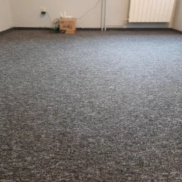 Szary dywan w pokoju z białymi ścianami i grzejnikiem, w rogu karton z routerem.