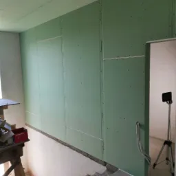 Ściana wewnętrzna z płyt kartonowo-gipsowych, widoczne łączenia, częściowo wykończona, z przewodami elektrycznymi wychodzącymi przez otwór. W tle widoczny statyw z lampą.