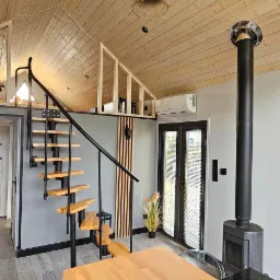 Wnętrze domu z drewnianym stropem, schodami z metalową balustradą, piecem wolnostojącym i drewnianym stołem, oświetlone wiszącą lampą z metalowym abażurem.