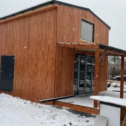 Dwukondygnacyjny dom z drewnianą elewacją w trakcie budowy, częściowo wykończony, z zadaszonym tarasem, otoczony śniegiem i materiałami budowlanymi.