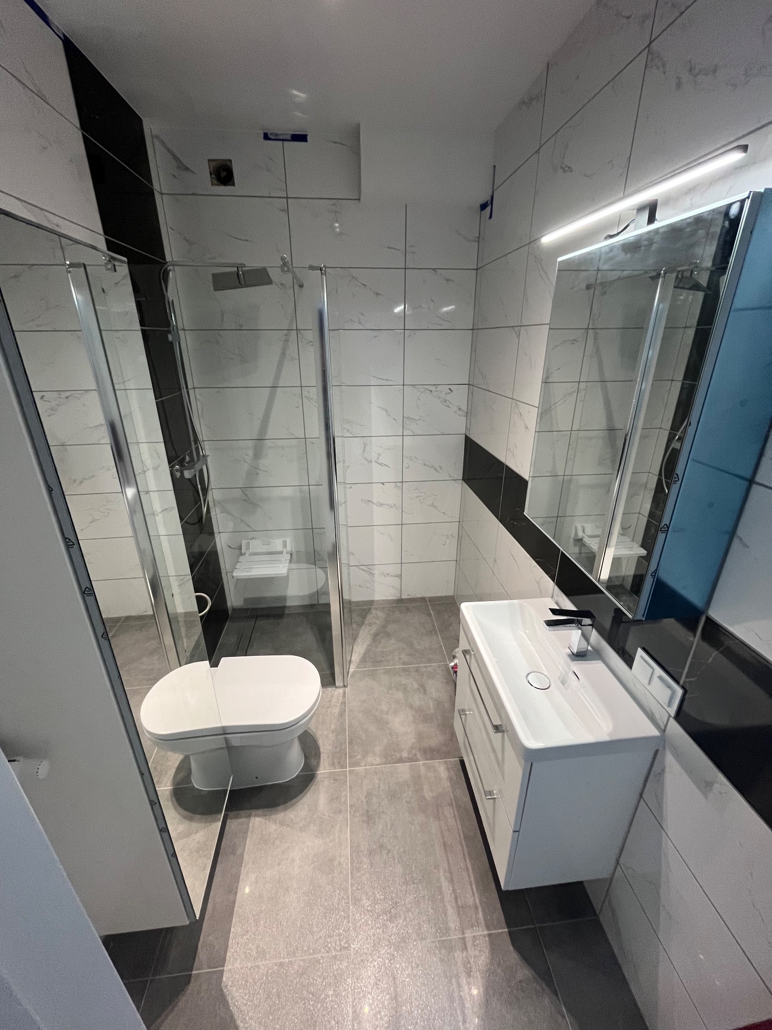 Nowoczesna łazienka z kabiną prysznicową ze szklanymi drzwiami, wiszącą toaletą, białą szafką z umywalką i podświetlanym lustrem, wykończona szaro-białymi płytkami z czarnymi akcentami.