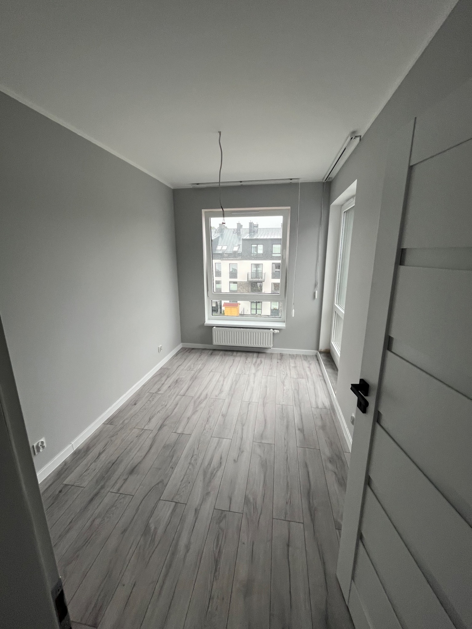 Pomalowane na szaro wnętrze pokoju z panelami podłogowymi imitującymi drewno, widok na budynki przez okno, białe drzwi z czarną klamką po prawej.