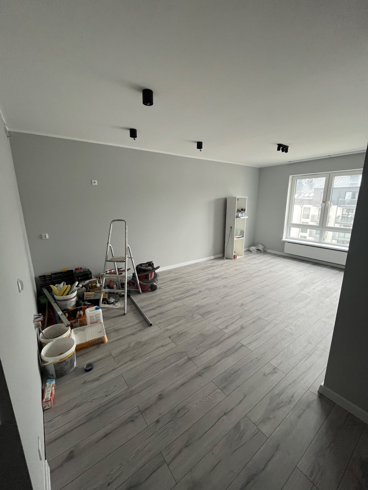Pokój w trakcie prac wykończeniowych: szare ściany, panele podłogowe imitujące drewno, drabina, narzędzia malarskie i wiaderka z farbą zgromadzone przy ścianie, widoczne lampy sufitowe i okno...