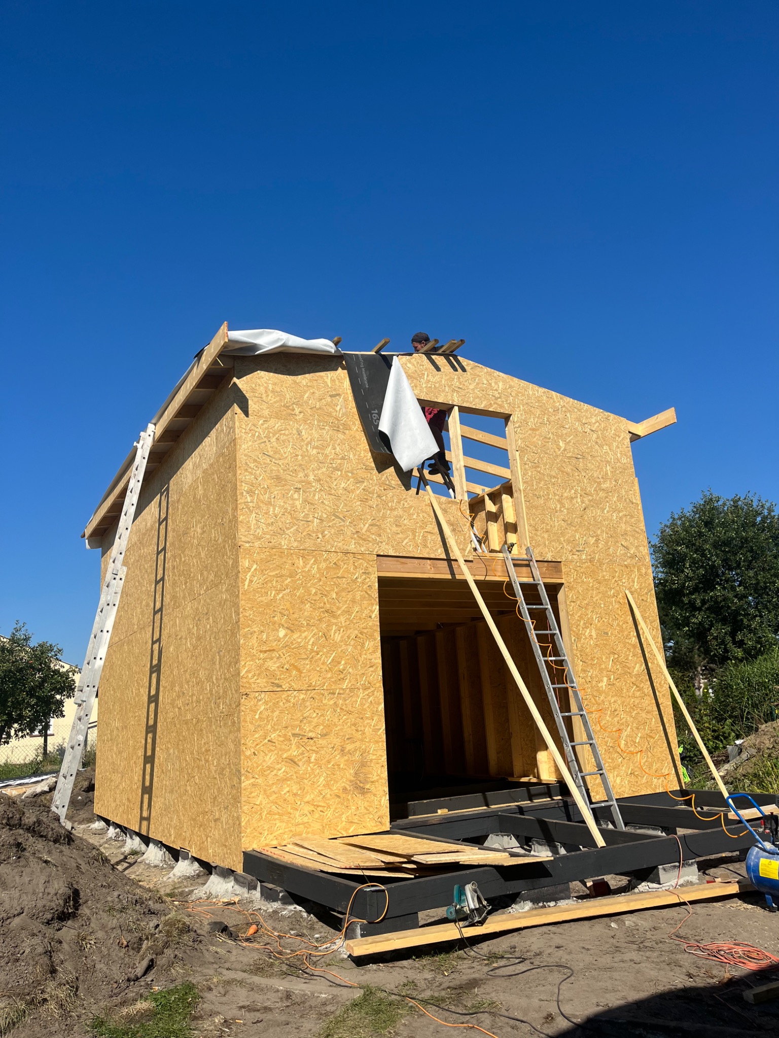 Budowa domu szkieletowego w Gryfinie: konstrukcja z płyty OSB, widoczny pracownik na dachu, drabiny oparta o ścianę, fundamenty i podkonstrukcja, błękitne niebo w tle.
