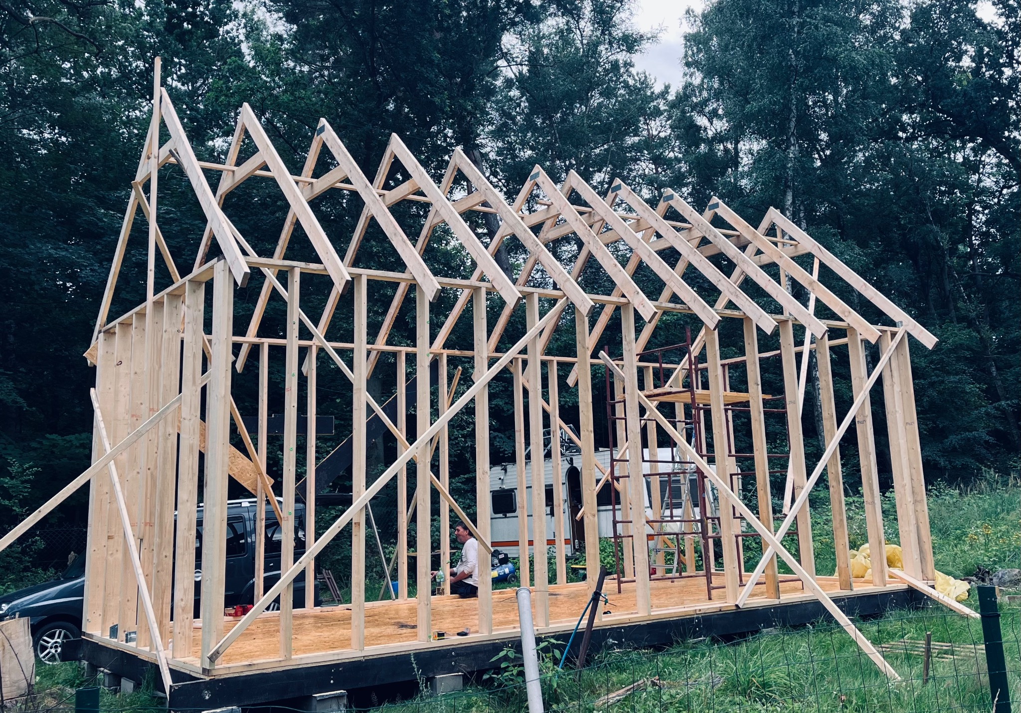 Wznoszenie konstrukcji szkieletowej domu z widocznym szkieletem dachu, pracownik siedzący wewnątrz, samochód i przyczepa kempingowa w tle na zewnątrz budynku.