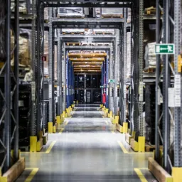 Długi korytarz pomiędzy regałami w magazynie, z żółtymi oznaczeniami podłogowymi i widocznym znakiem ewakuacyjnym.