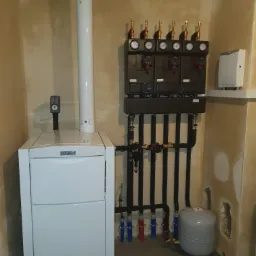 Biały piec grzewczy Vaillant z podłączonymi czarnymi rurami, manometry i zawory na ścianie w surowym pomieszczeniu kotłowni.