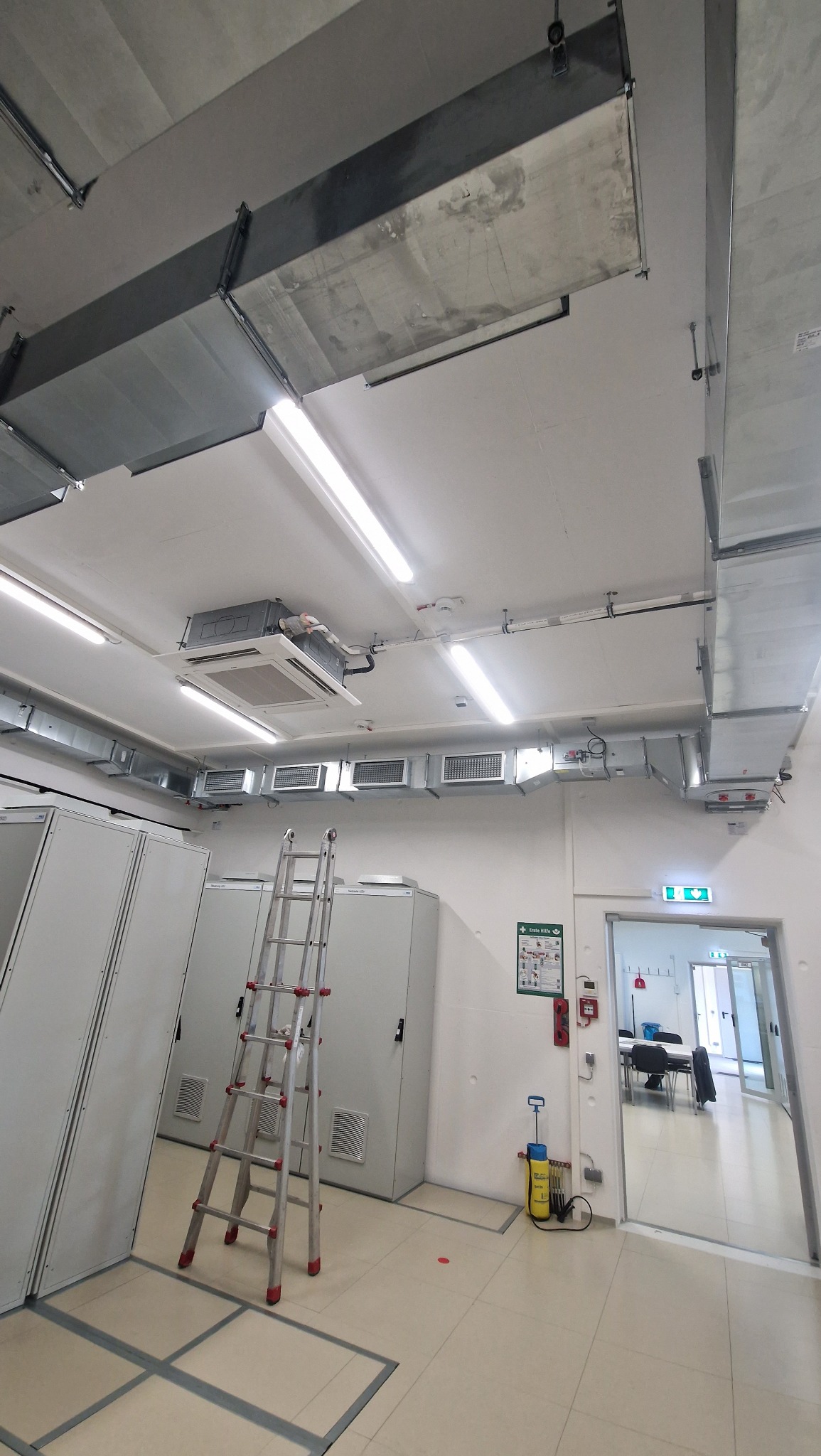 Instalacja wentylacji i klimatyzacji w pomieszczeniu technicznym z drabiną, widoczne kanały wentylacyjne, klimatyzator kasetonowy i szafy elektryczne.