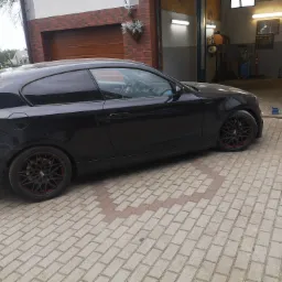 Czarne auto z felgami z czerwonym akcentem stoi przed warsztatem, obok leży skrzynia biegów.