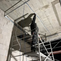 Olsan Andrzej Raduszyński - Elektryk na rusztowaniu montuje kable w metalowych prowadnicach pod surowym betonowym sufitem w budynku w stanie deweloperskim.