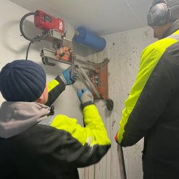 Olsan Andrzej Raduszyński - Dwóch pracowników wierci otwór w betonowej ścianie za pomocą wiertnicy diamentowej z odciągiem, widoczne ślady po wodzie.