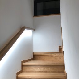 Olsan Andrzej Raduszyński - Drewniane schody z podświetlaną poręczą LED na białej ścianie, widok z dołu, nowoczesny design.
