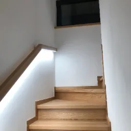 Drewniane schody z podświetlaną poręczą LED na białej ścianie, widok z dołu, nowoczesny design.