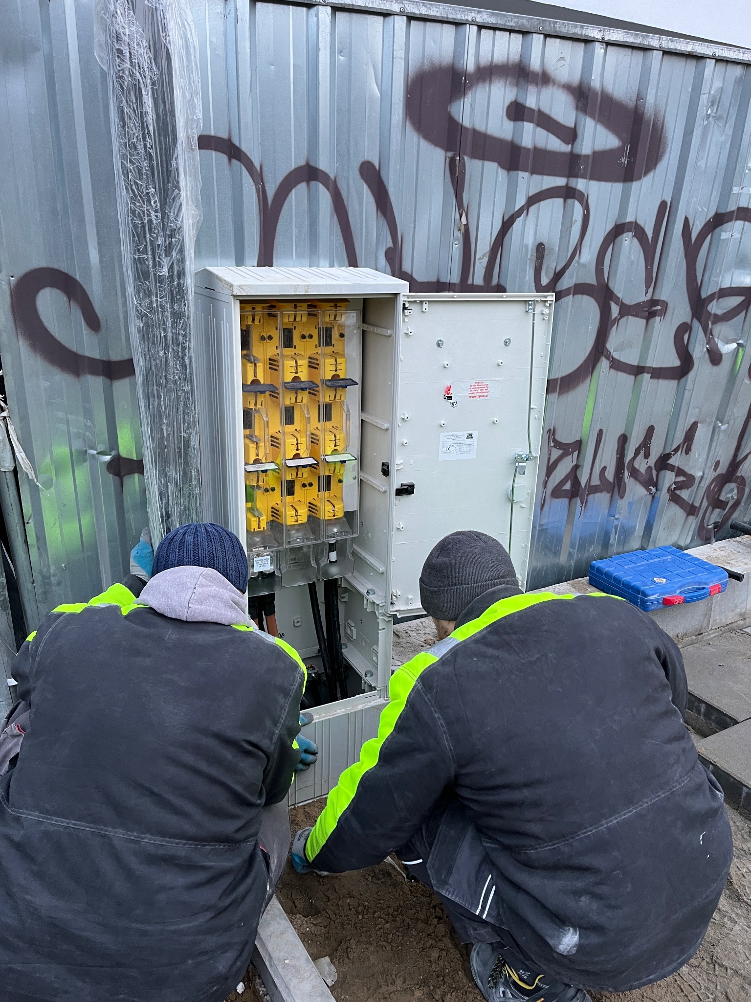 Dwóch elektryków w ubraniach roboczych z elementami odblaskowymi pracuje przy otwartej skrzynce z bezpiecznikami na tle blaszanej ściany pokrytej graffiti.