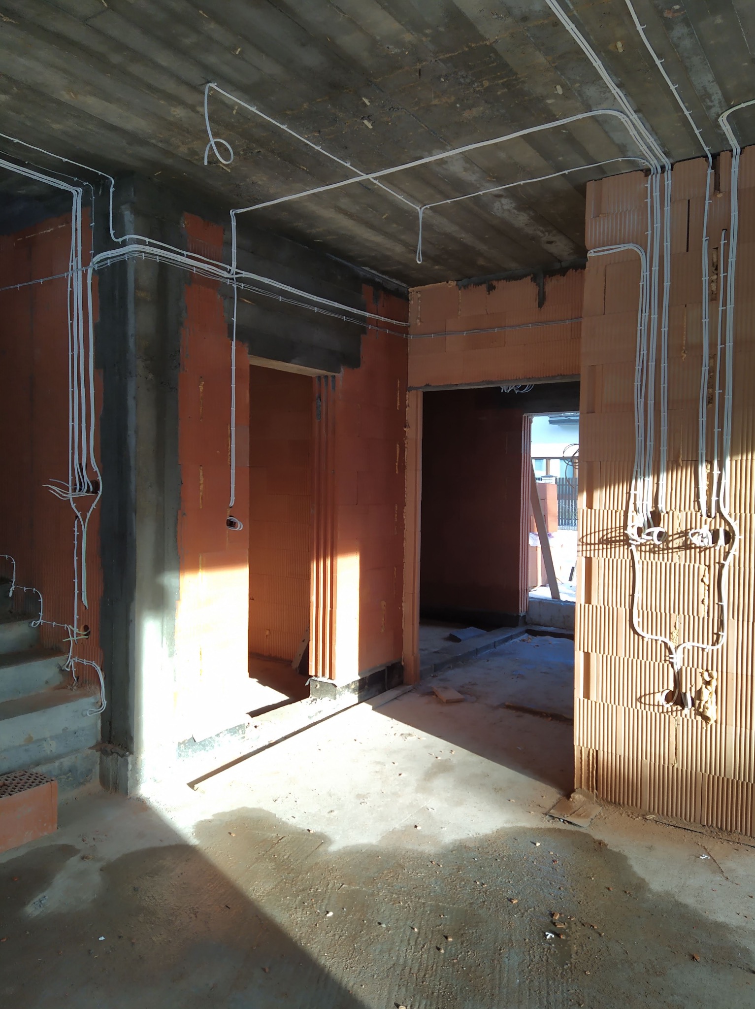 Rozprowadzone kable elektryczne w białych peszlach na ścianach z pustaków ceramicznych i suficie surowego, betonowego budynku w trakcie budowy, widoczne otwory drzwiowe i fragment schodów.