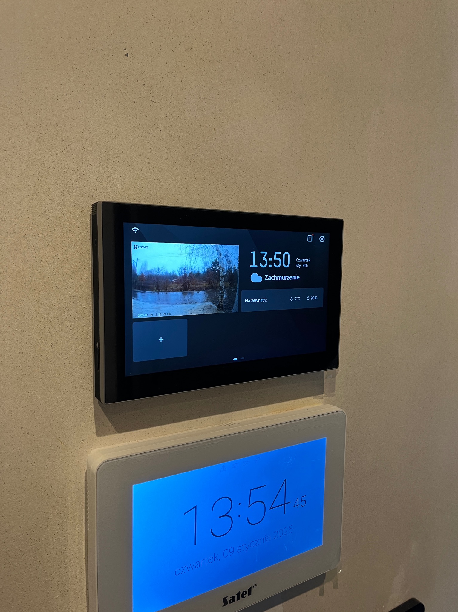 Zamontowany na ścianie panel sterowania systemem monitoringu wizyjnego, wyświetlający obraz z kamery zewnętrznej, datę, godzinę i temperaturę, umieszczony nad panelem alarmowym Satel.