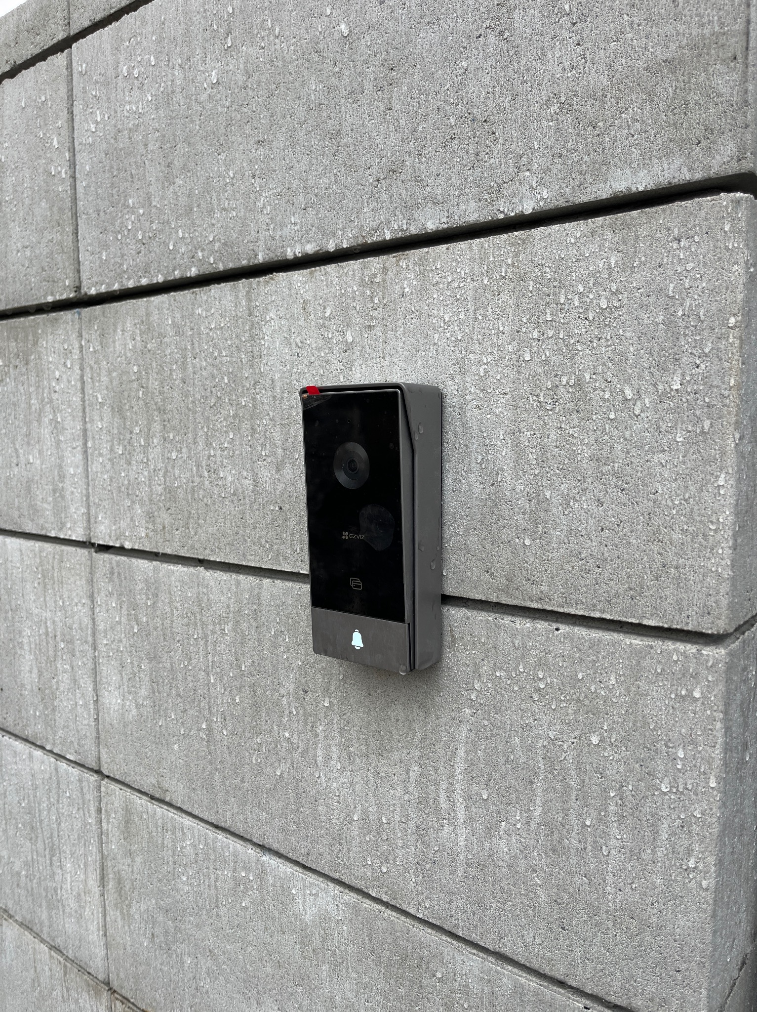 Czarny wideodomofon marki EZVIZ zamontowany na szarej, betonowej ścianie z widocznymi kroplami deszczu.
