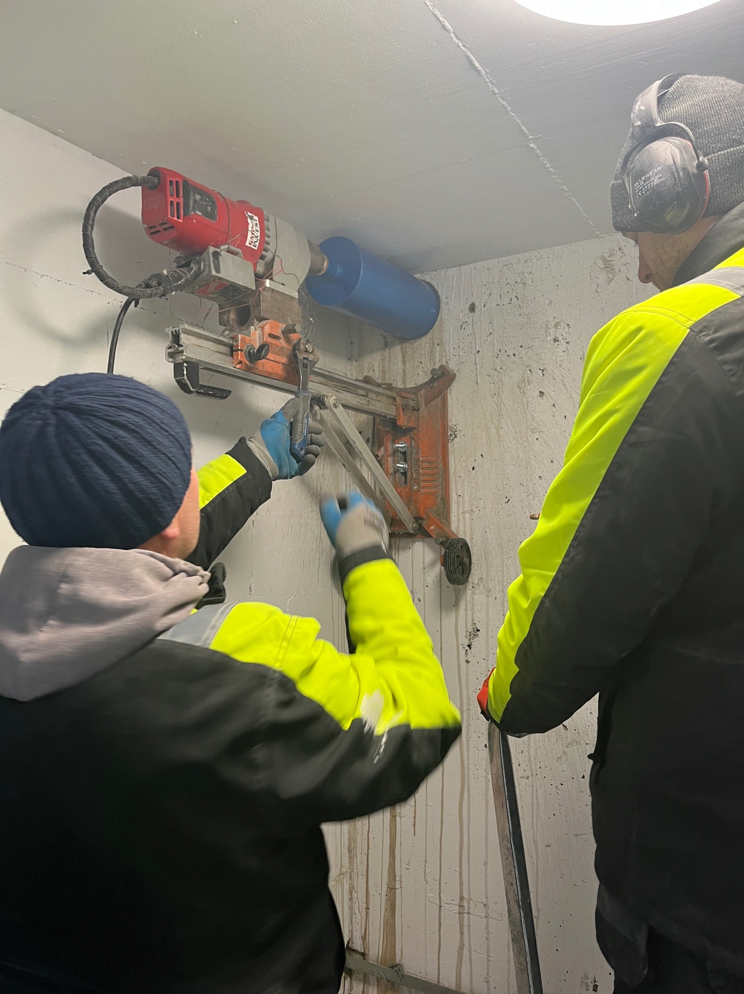 Dwóch pracowników wierci otwór w betonowej ścianie za pomocą wiertnicy diamentowej z odciągiem, widoczne ślady po wodzie.