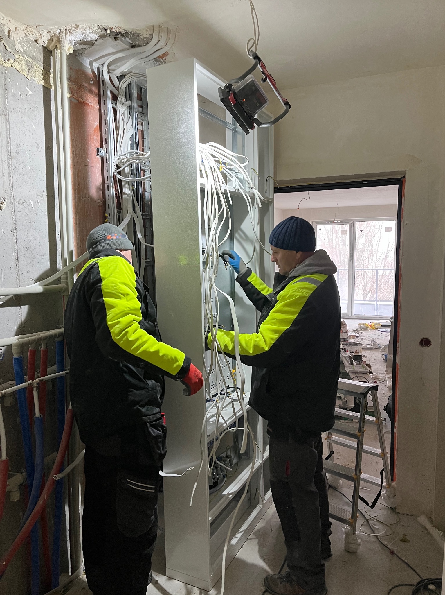Dwóch elektryków w Słupnie montuje instalację elektryczną w nowym budynku, widoczna skrzynka rozdzielcza z licznymi kablami, oświetlenie robocze i drabina.