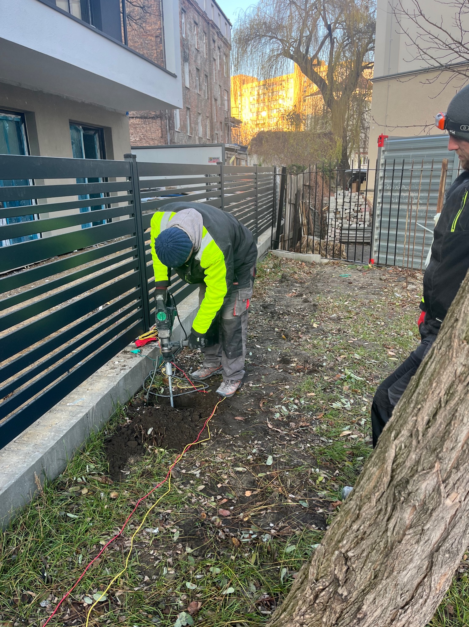 Pracownik w odblaskowej kamizelce wierci otwór w ziemi w pobliżu ogrodzenia, prawdopodobnie pod uziom instalacji odgromowej, widoczne przewody i narzędzia.