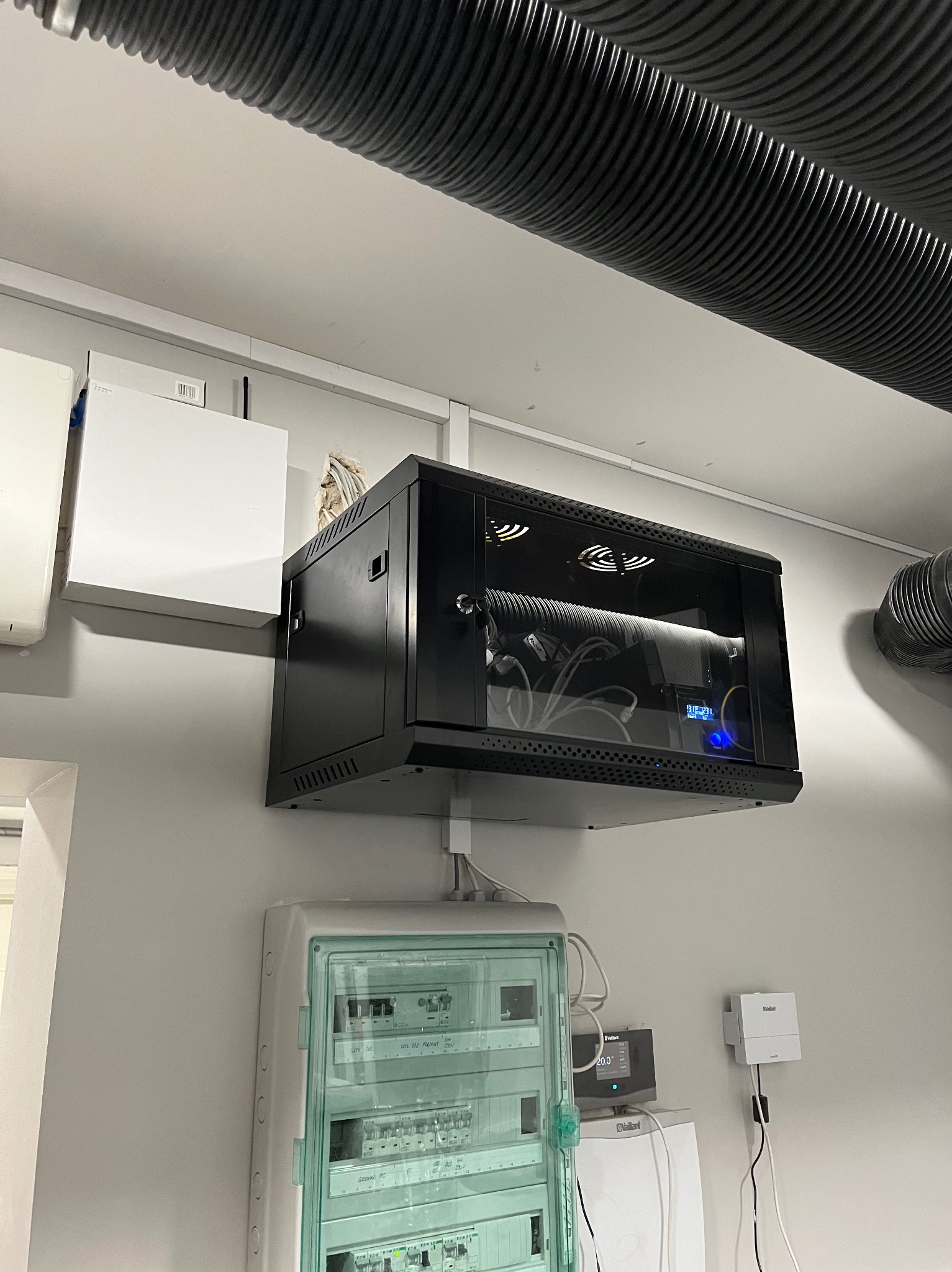 Szafa rack wisząca na ścianie, z widocznymi kablami i urządzeniami wewnątrz, poniżej rozdzielnica elektryczna w obudowie z zielonymi akcentami, na tle białej ściany i czarnej rury wentylacyjnej...