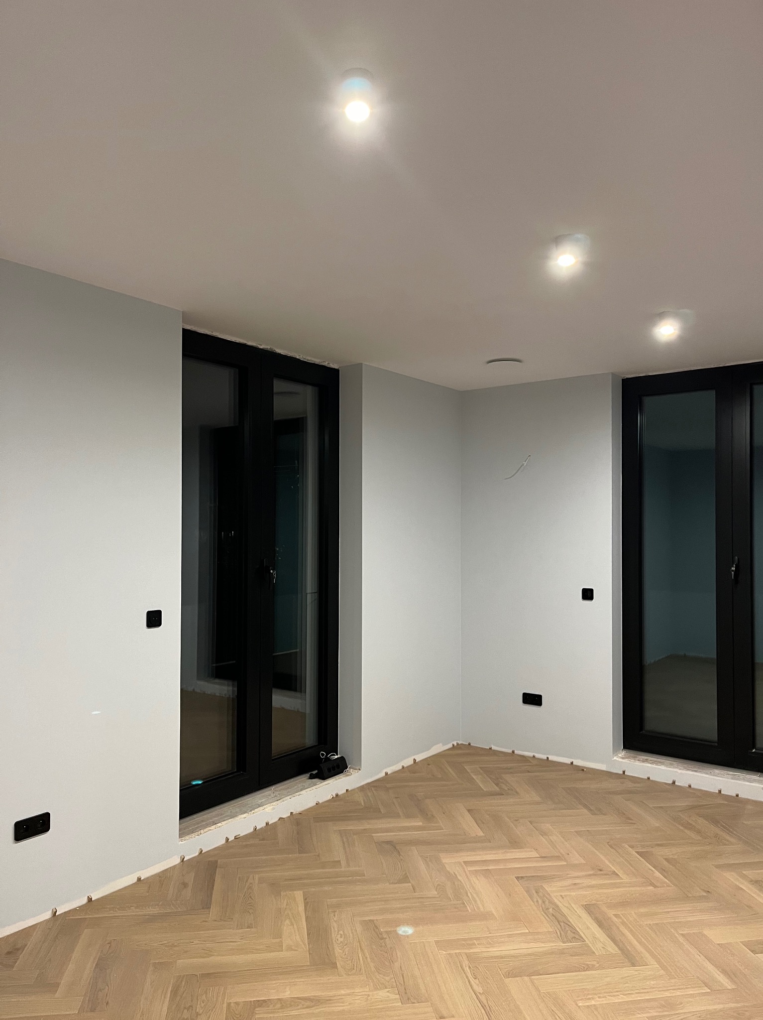 Nowoczesne wnętrze z parkietem ułożonym w jodełkę, czarne ramy okienne i gniazdka elektryczne, białe ściany z widocznymi otworami po instalacji elektrycznej i punktowe oświetlenie sufitowe.