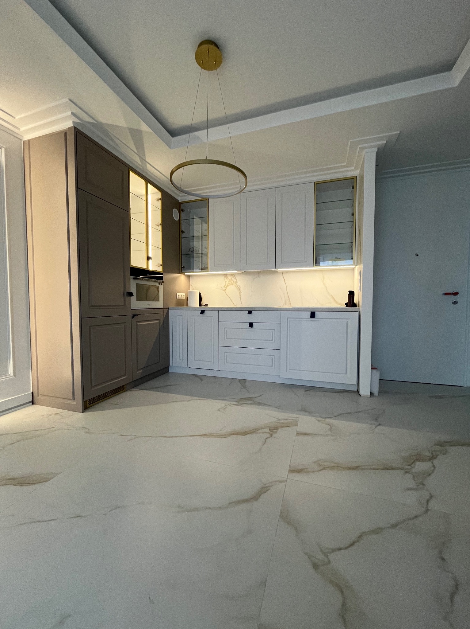 Nowoczesna kuchnia z białymi i szarymi szafkami, marmurowym blatem i złotymi akcentami, oświetleniem LED pod szafkami i okrągłą, złotą lampą wiszącą.