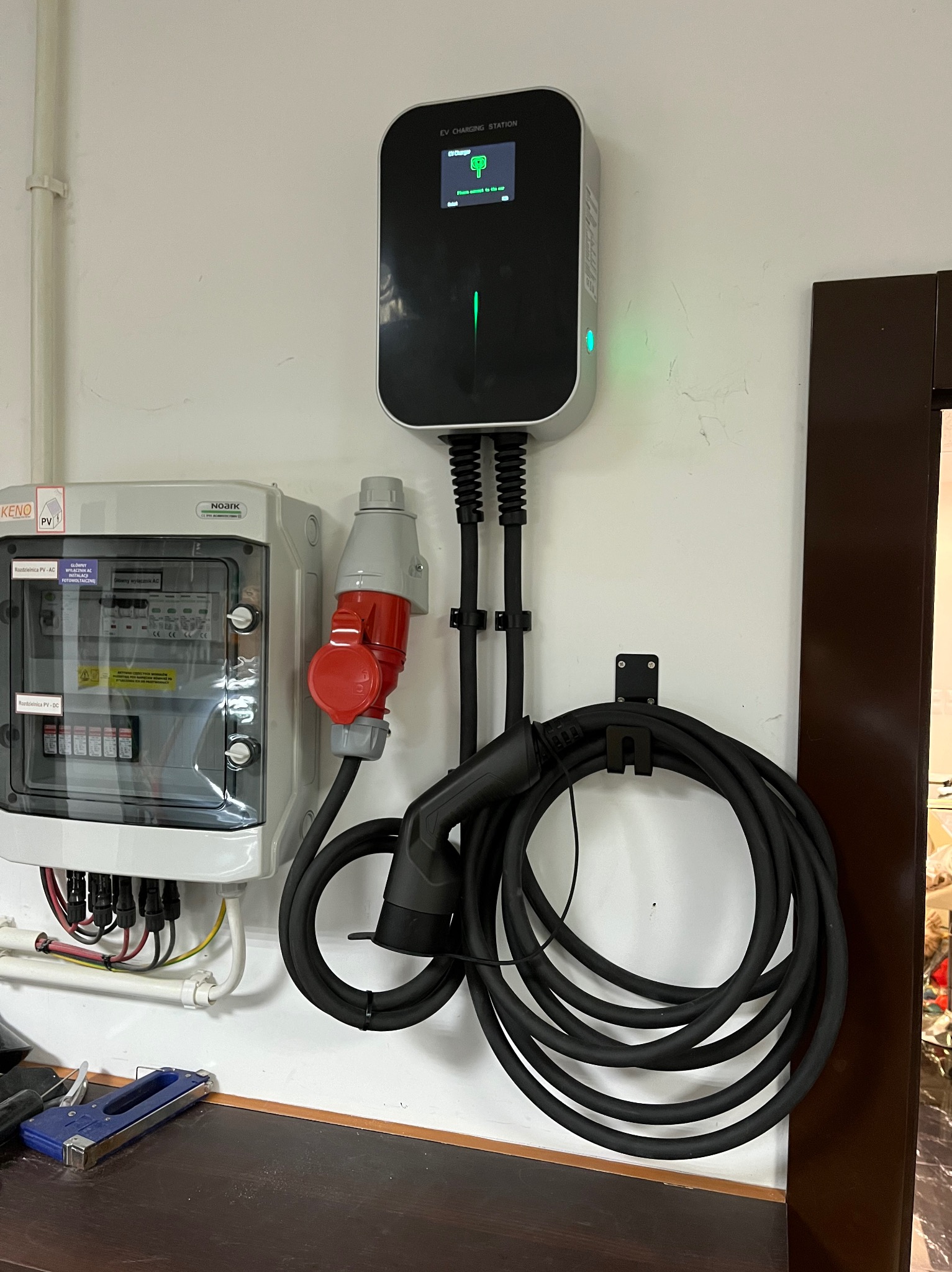 Stacja ładowania pojazdów elektrycznych zamontowana na ścianie, obok rozdzielnicy PV z czerwonym gniazdem siłowym, kabel zwinięty na uchwycie, na blacie zszywacz tapicerski.