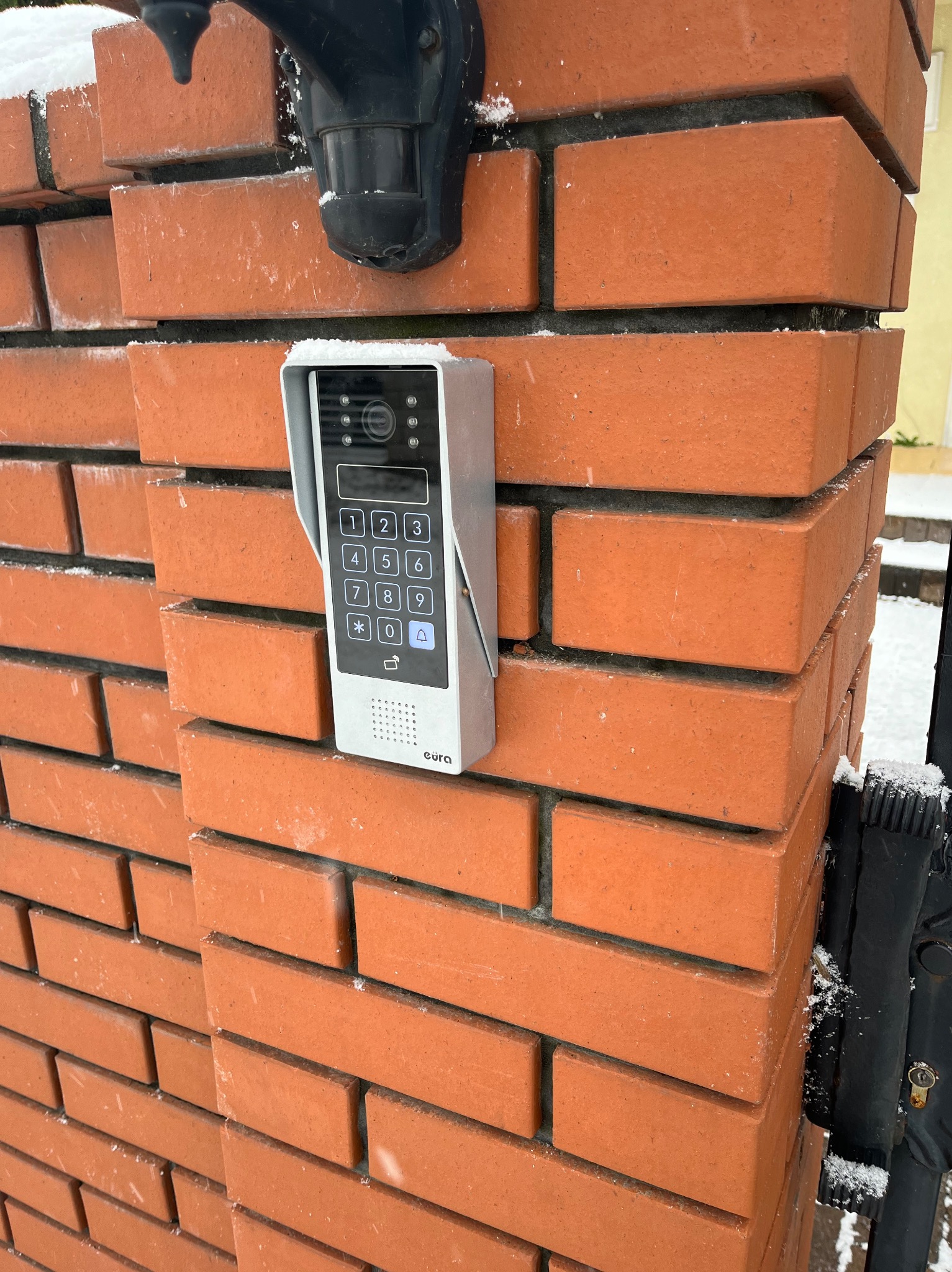 Zamontowany wideofon Eura z klawiaturą numeryczną na ceglanym słupku ogrodzenia w Słupnie, widoczne ślady śniegu.