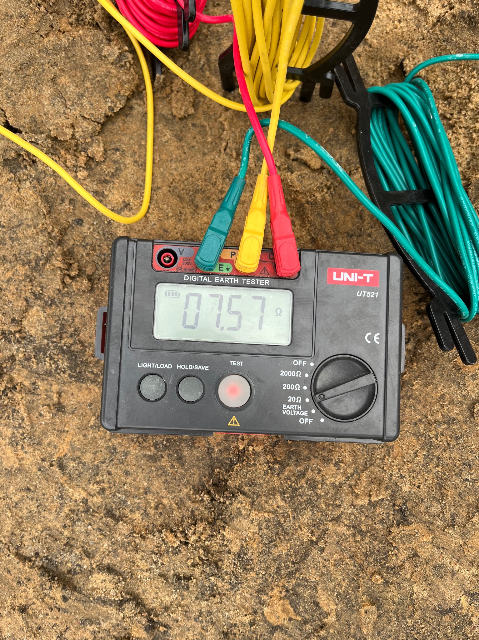 Cyfrowy miernik uziemiający UNI-T UT521 z podłączonymi przewodami pomiarowymi w kolorach czerwonym, żółtym i zielonym, leżący na piaszczystym podłożu.