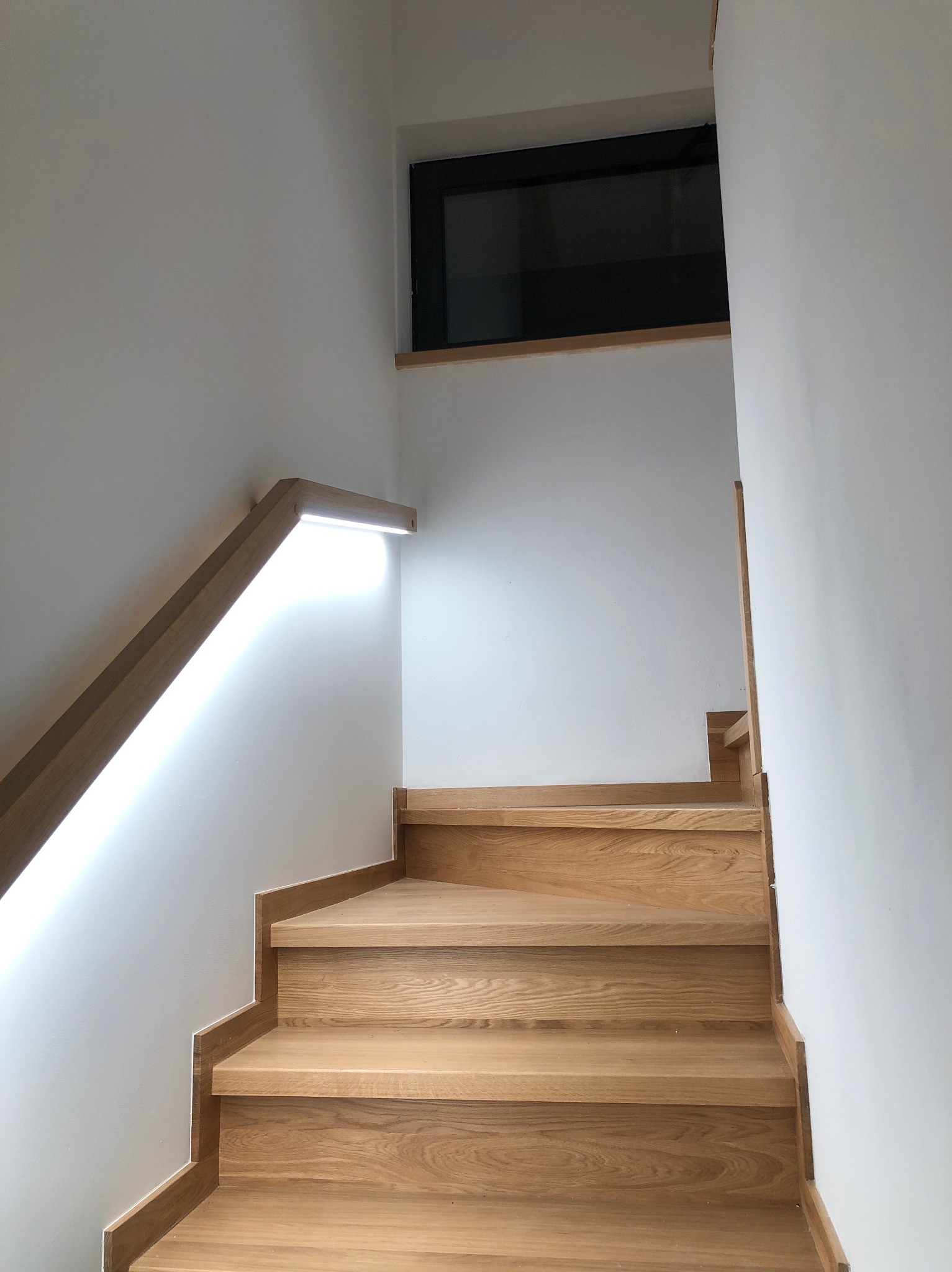 Drewniane schody z podświetlaną poręczą LED na białej ścianie, widok z dołu, nowoczesny design.