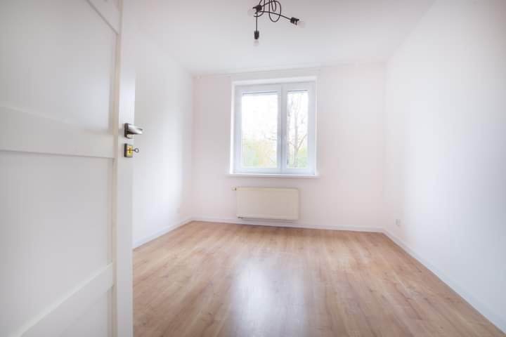 Pusty, jasny pokój z drewnianą podłogą, białymi ścianami i oknem z widokiem na drzewa, otwarte białe drzwi z widoczną klamką po lewej stronie.