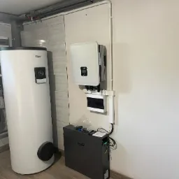 Biały inwerter fotowoltaiczny H2O, akumulator Fox-Ess i zbiornik Valami zamontowane na ścianie w pomieszczeniu z brązową podłogą imitującą drewno.