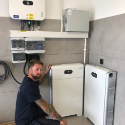 Sławomir Wasilik Voltum Kompleksowe Usługi Elektryczne - Instalacja fotowoltaiczna w pomieszczeniu: biały inwerter Huawei, akumulatory energii i rozdzielnia. Instalator z tatuażami przy akumulatorach.