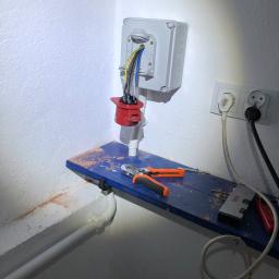 Sławomir Wasilik Voltum Kompleksowe Usługi Elektryczne - Szara puszka elektryczna z wystającymi przewodami, podłączona do czerwonego gniazda przemysłowego na ścianie, narzędzia i wkręty na niebieskiej półce, gniazdko elektryczne obok.