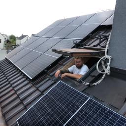 Sławomir Wasilik Voltum Kompleksowe Usługi Elektryczne - Mężczyzna z brodą wystaje z okna dachowego na dachu z panelami fotowoltaicznymi, widoczne kable i fragmenty instalacji.